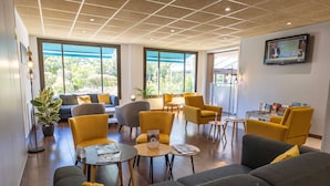 A modern lounge area with sofas, chairs, and tables, large windows with a view, and a flat-screen TV on the wall. - Kyriad Blois Sud-Vineuil (Vineuil)