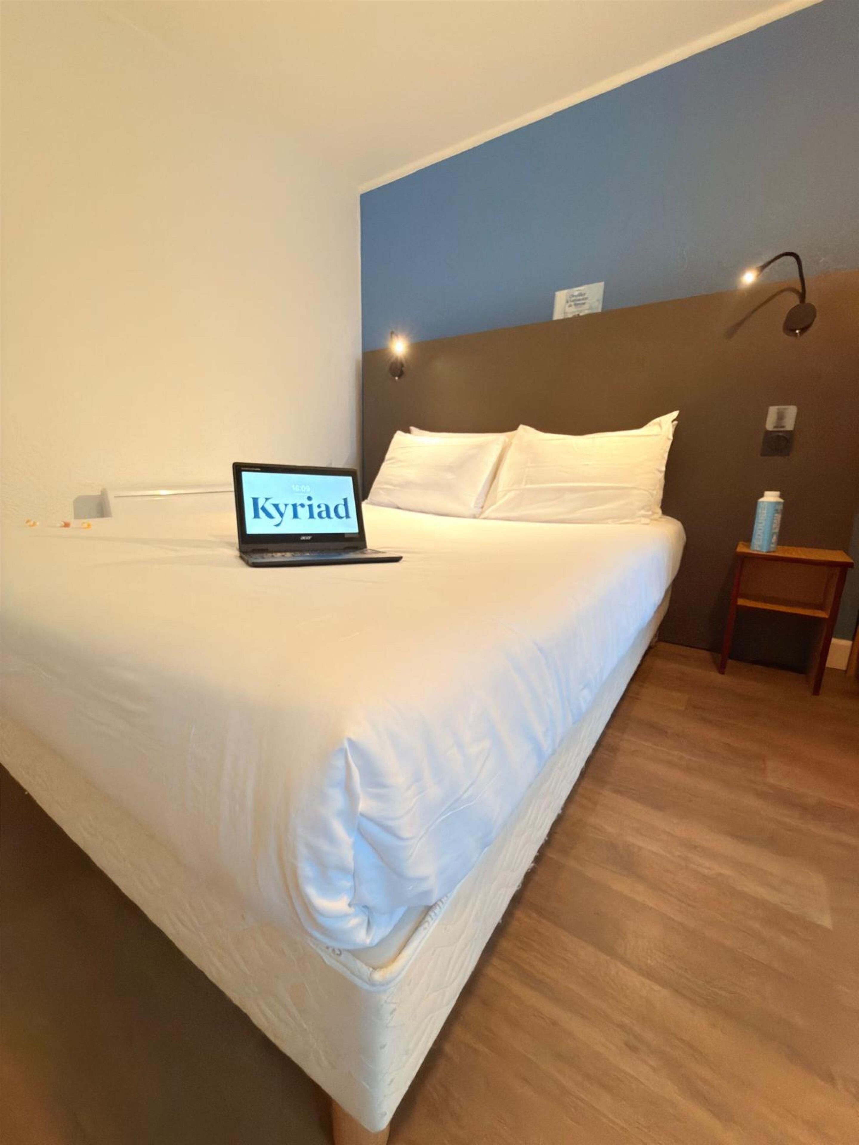 Une chambre d’hôtel avec un lit, un ordinateur portable posé sur le lit, une table de chevet avec une bouteille et une lampe fixée au mur.