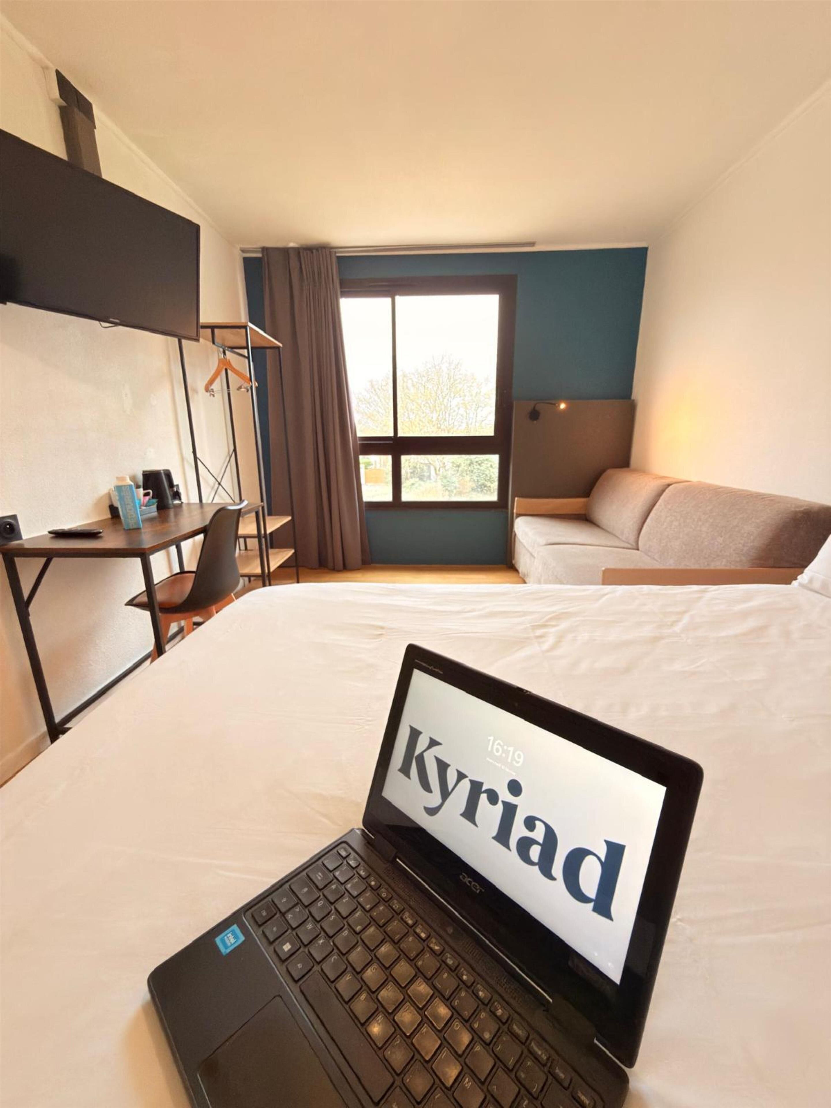Quarto de hotel com uma cama, uma escrivaninha com um laptop, uma cadeira, uma janela com cortinas e um sofá.