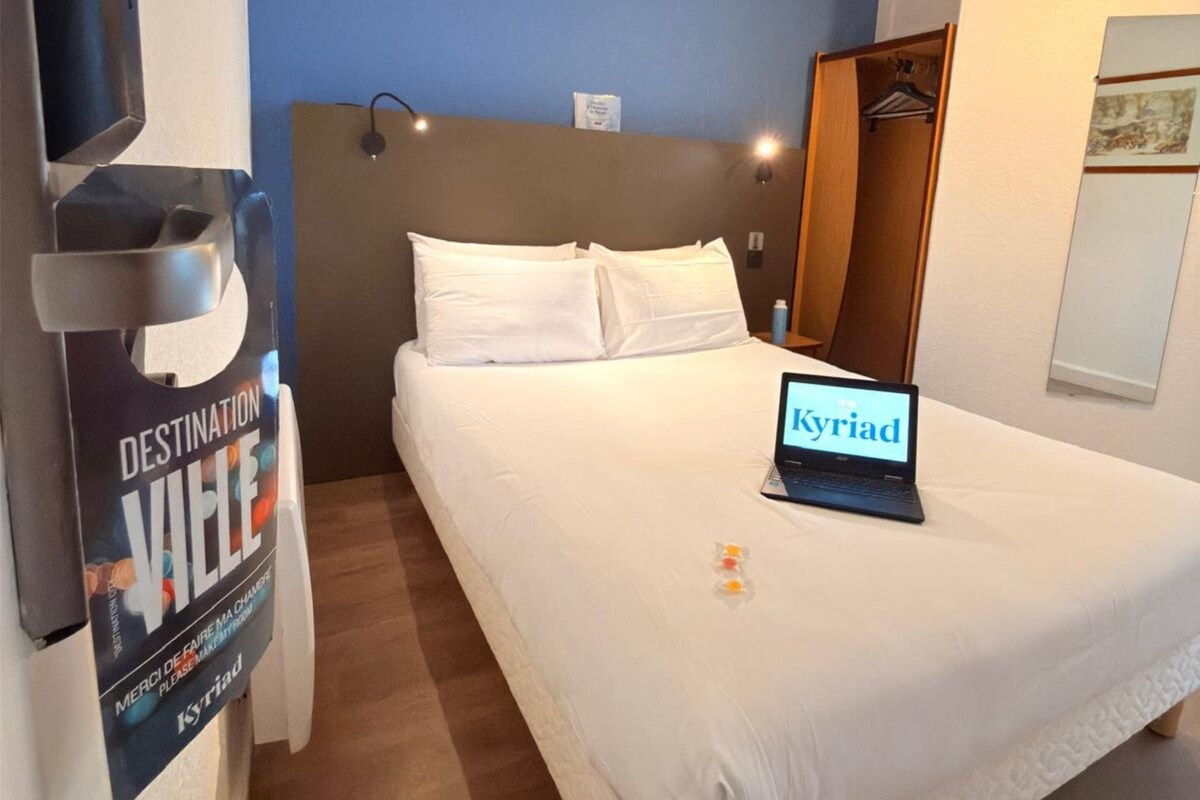 A hotel room with a bed, a laptop displaying 'Kyriad', a poster on the wall, and a wooden floor.