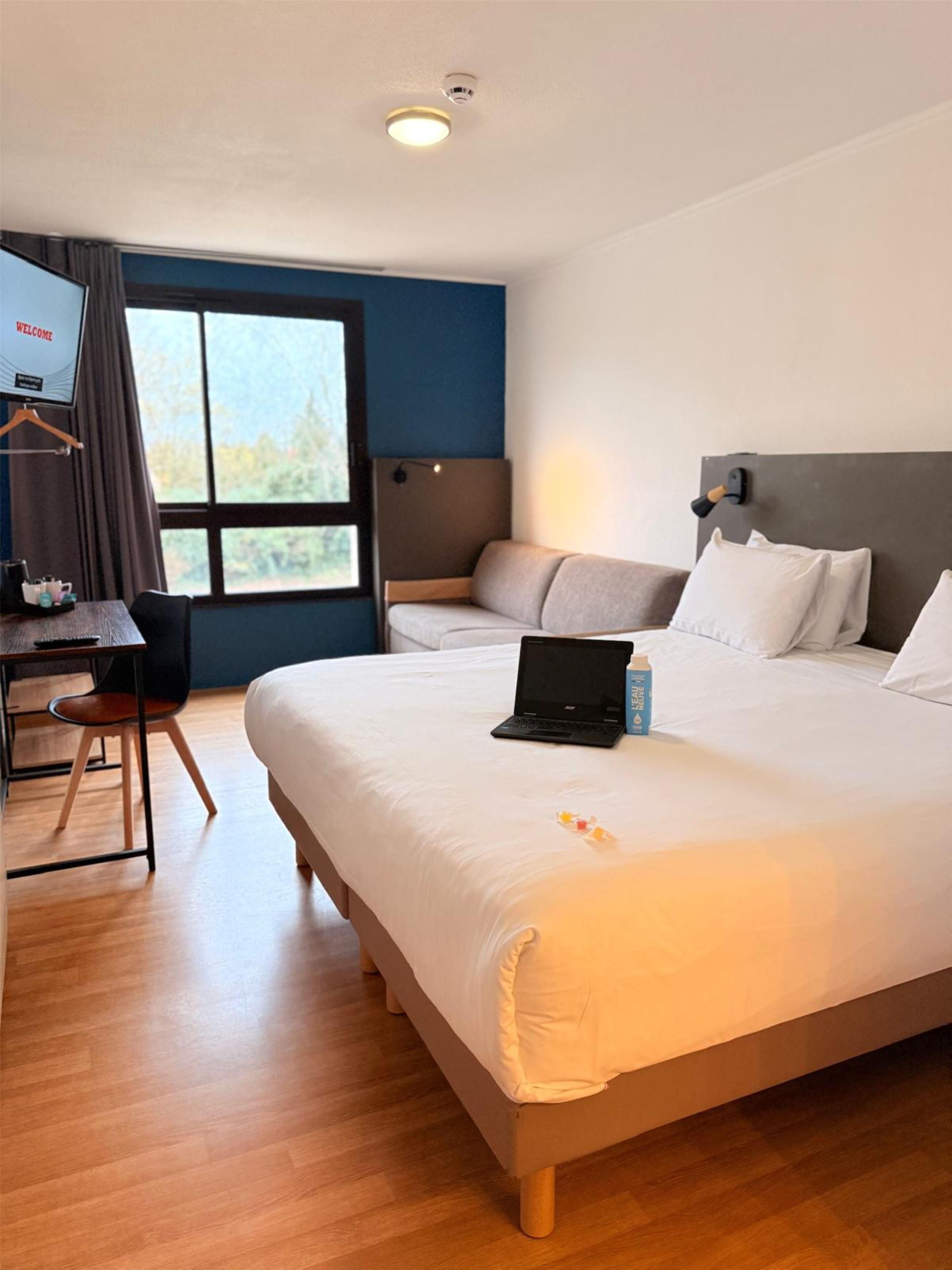 Habitación de hotel con cama, escritorio con una computadora portátil, silla, ventana con vista a árboles y televisor montado en la pared.