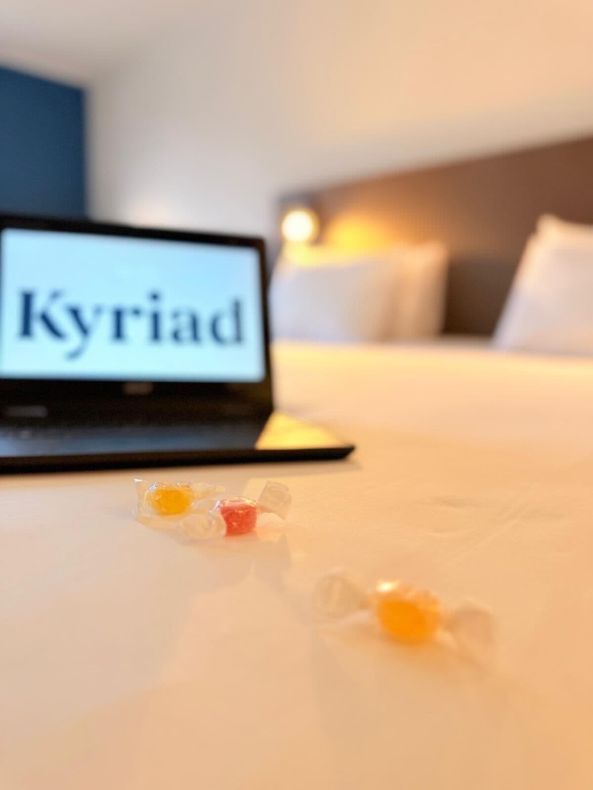 Un portatile con 'Kyriad' sullo schermo, un letto con biancheria bianca e un comodino con un piccolo oggetto.
