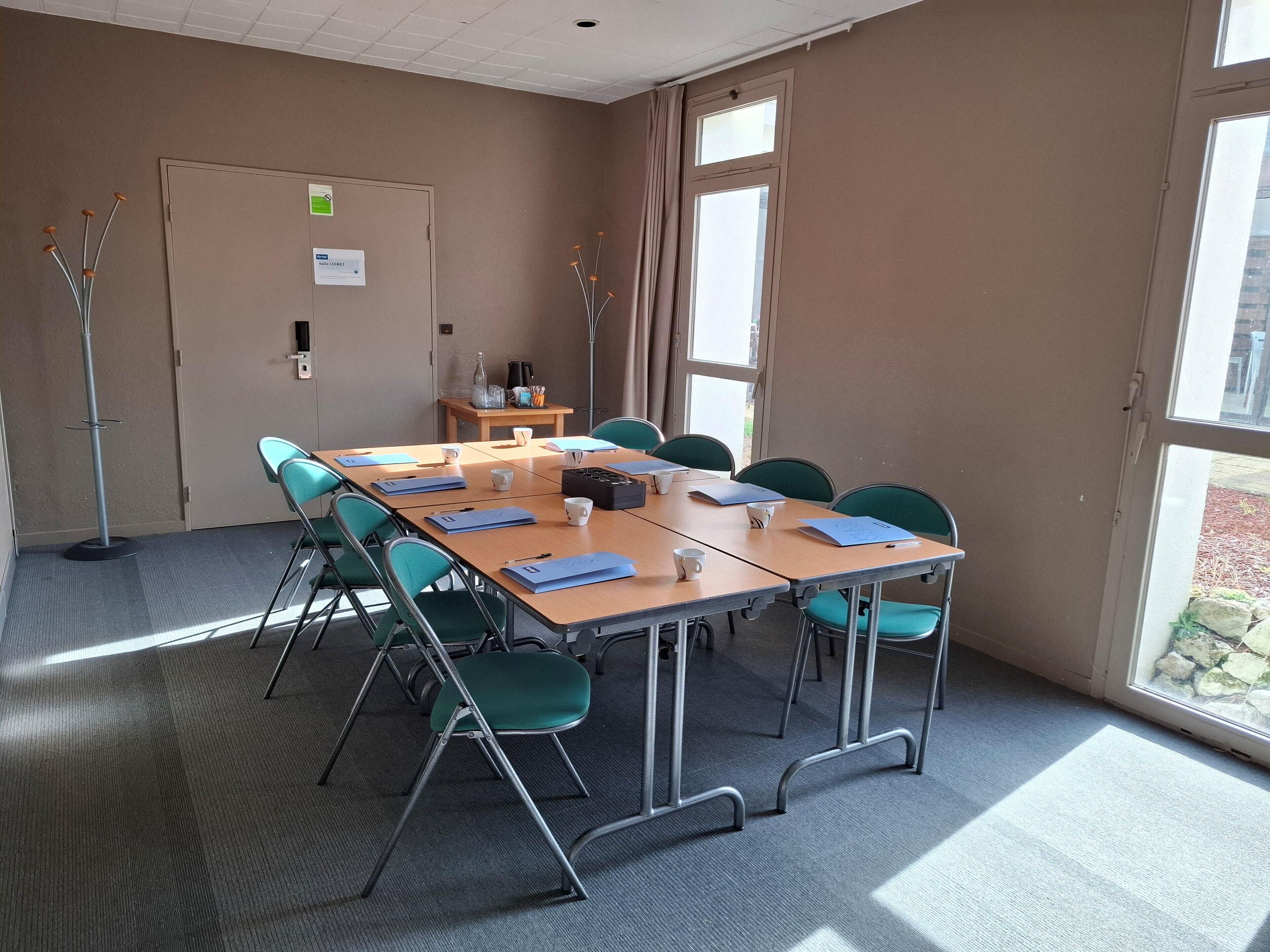 A conference room with a long table, green chairs, and a small table with a bottle and cups.