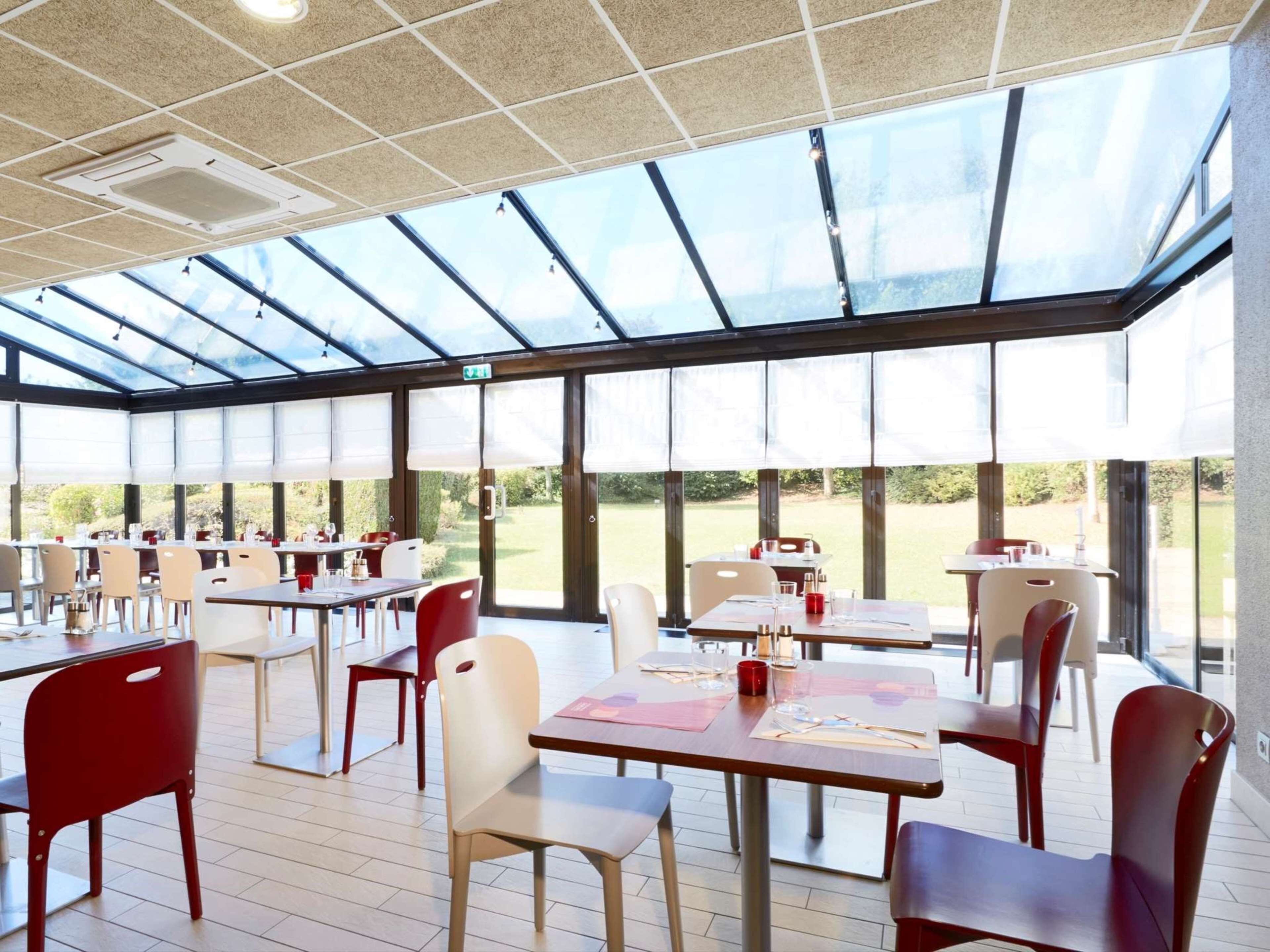 A spacious indoor dining area with tables and chairs, large windows, and a ceiling with a grid pattern.