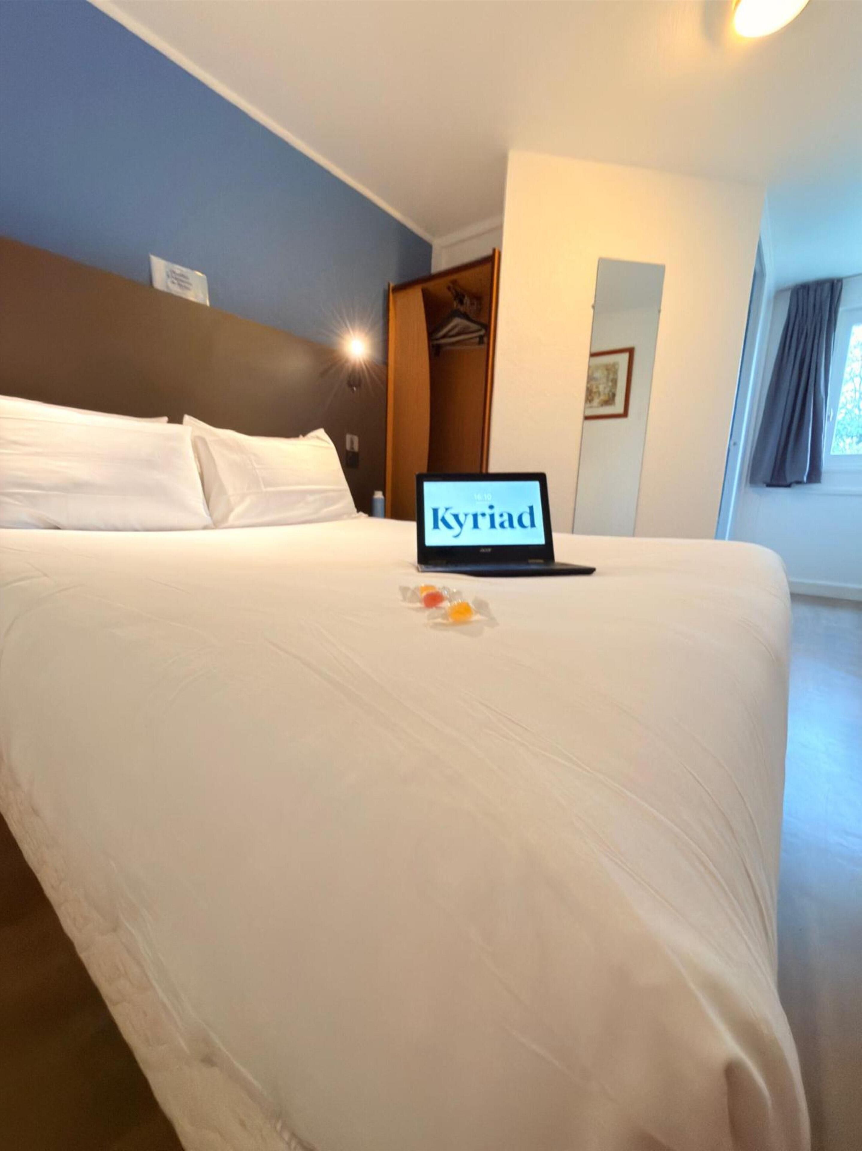 A hotel room with a bed, a laptop displaying 'Kyriad', a wardrobe, and a window with curtains.