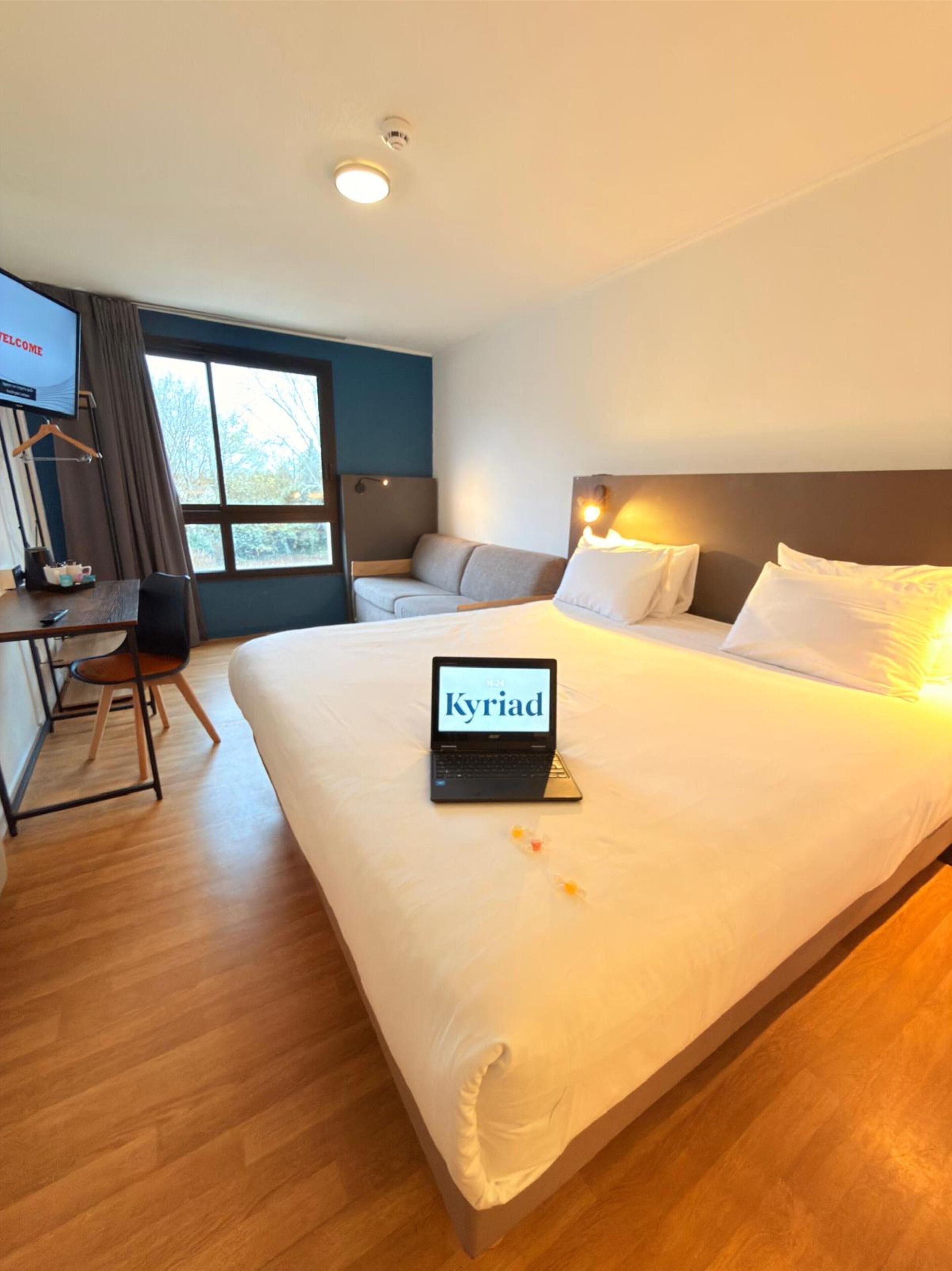 Une chambre d’hôtel avec un lit, un bureau avec un ordinateur portable, un canapé et un téléviseur fixé au mur.