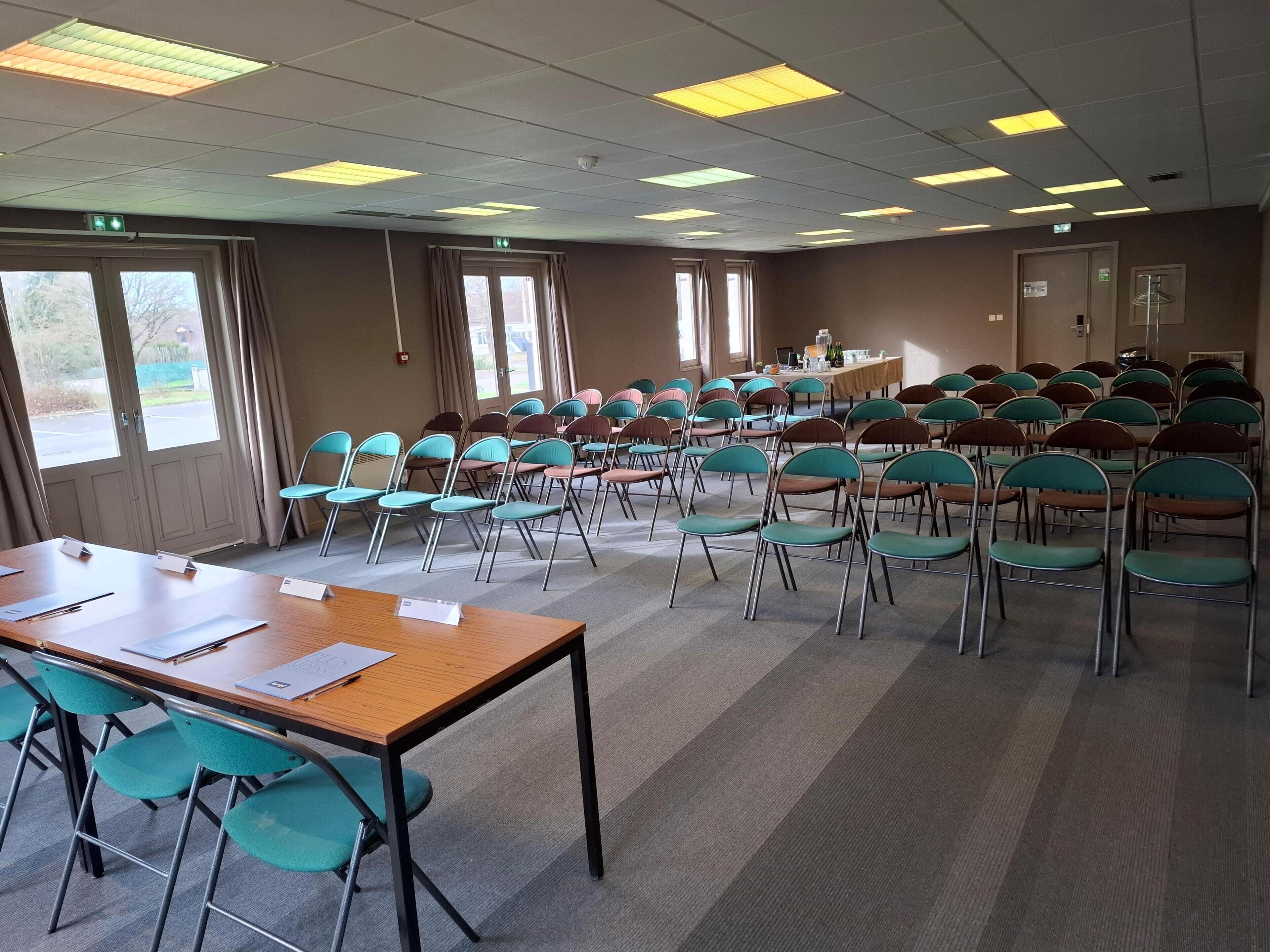 A conference room with rows of teal chairs, a long table with papers, and large windows allowing natural light.