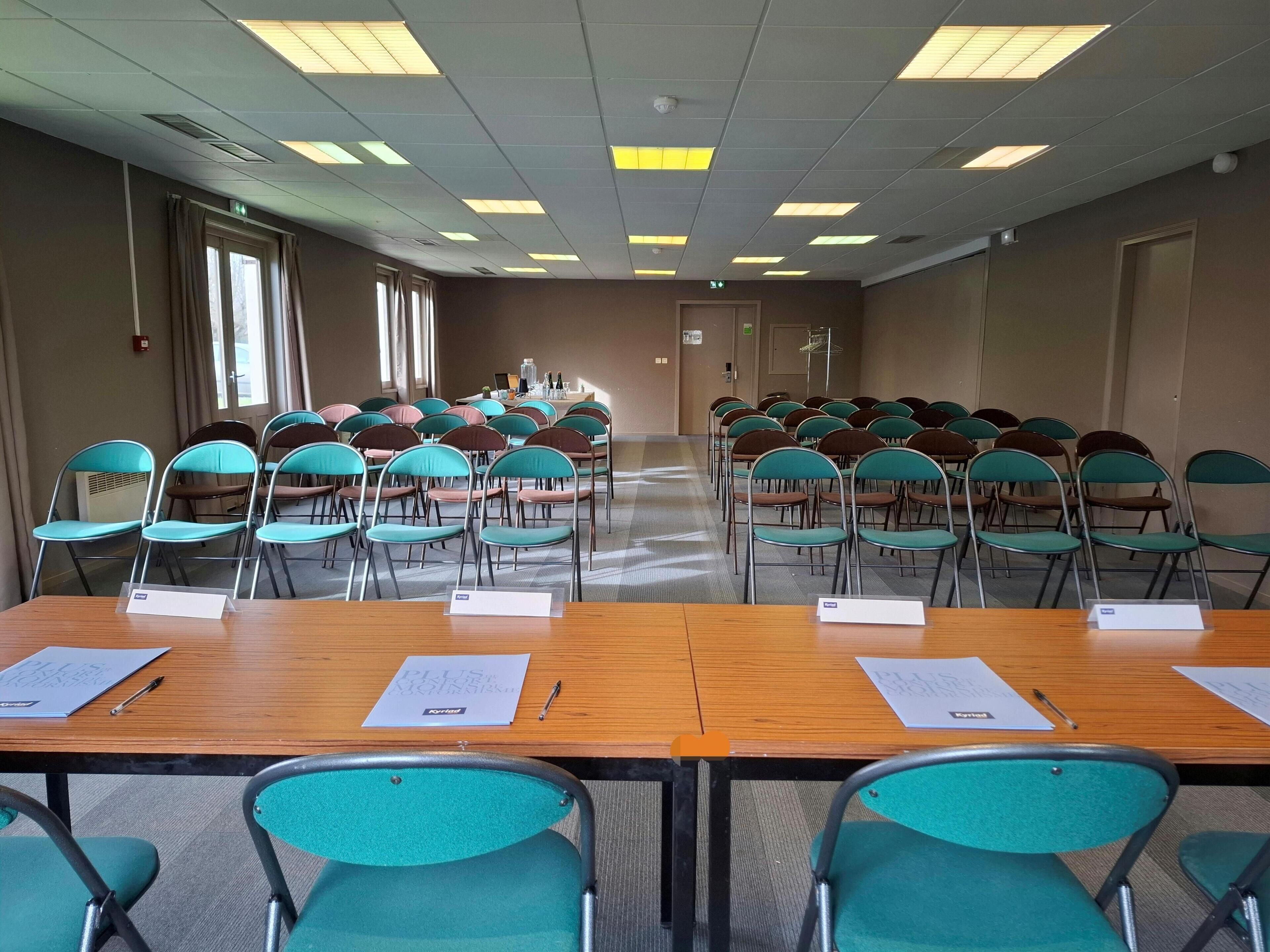 A conference room with rows of teal chairs, wooden tables with papers, and a ceiling with fluorescent lights.
