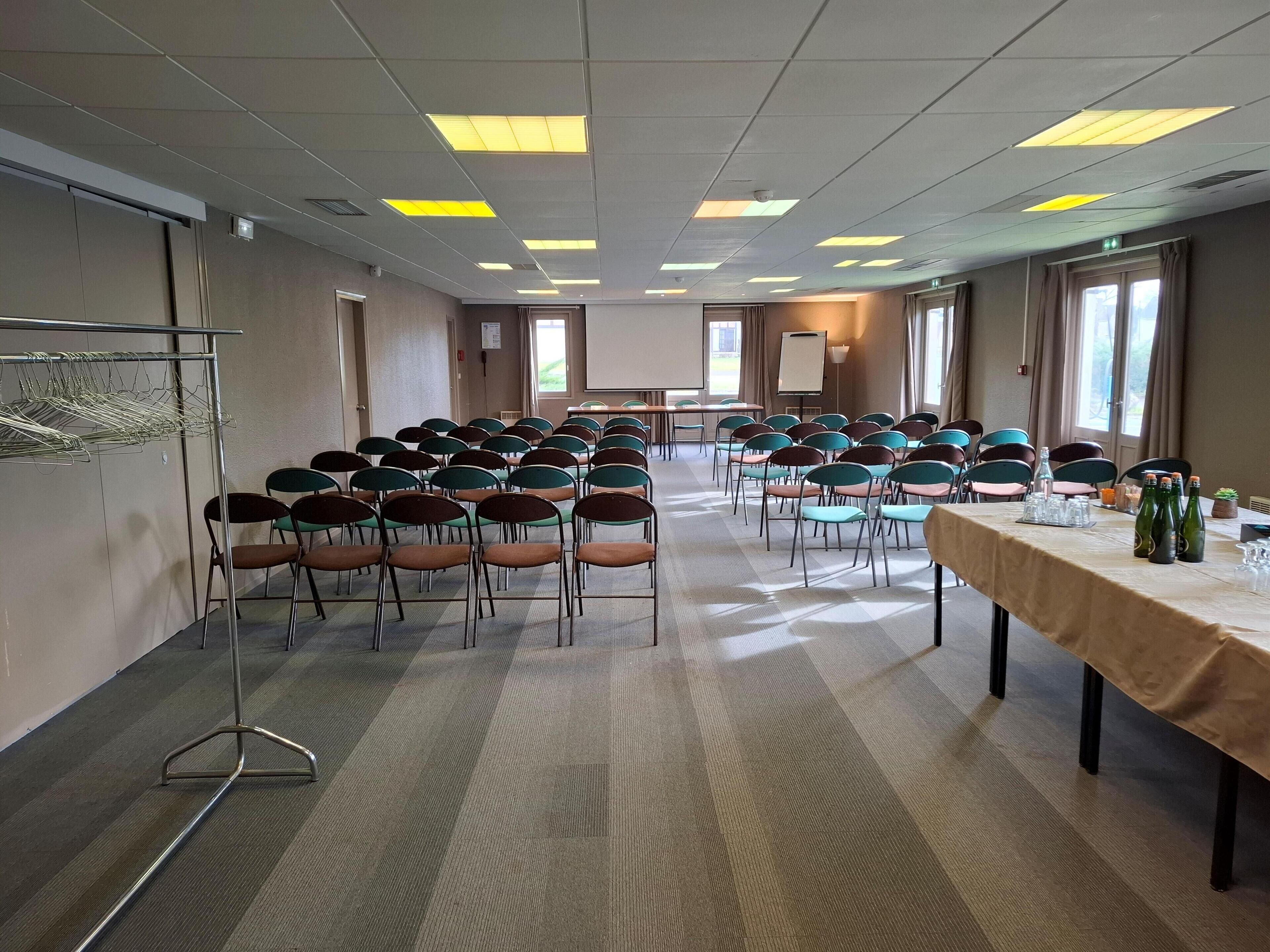 A conference room with rows of chairs, a long table with bottles and glasses, and a projector screen.
