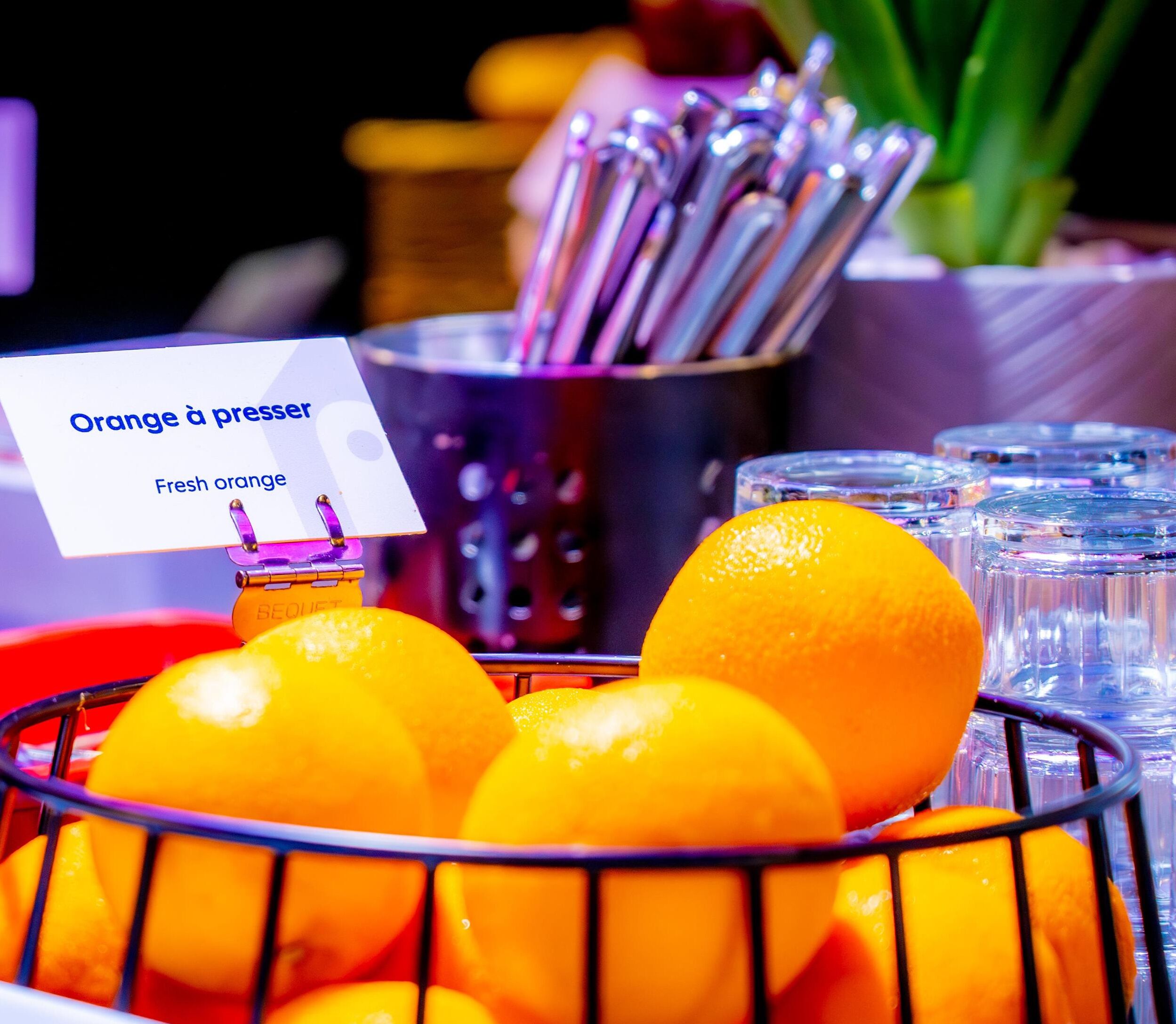 Una cesta de naranjas frescas con un cartel que dice 'Orange à presser' y un recipiente metálico con utensilios.