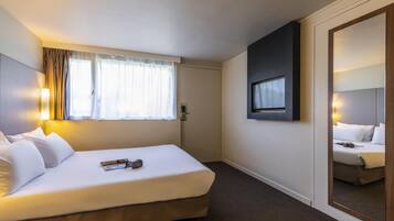 A hotel room with a bed, a window with curtains, a wall-mounted TV, and a mirror.