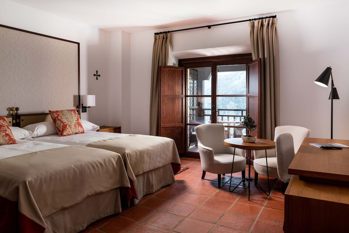 Habitación de hotel con dos camas, una mesita con sillas, un escritorio, una lámpara y un ventanal con cortinas.