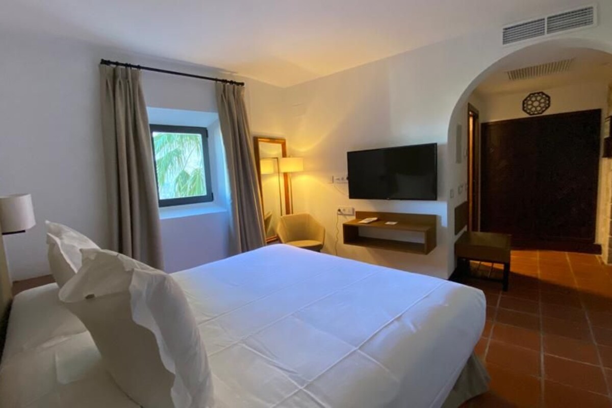 Habitación de hotel con una cama grande, un televisor montado en la pared, una silla y una ventana con cortinas.