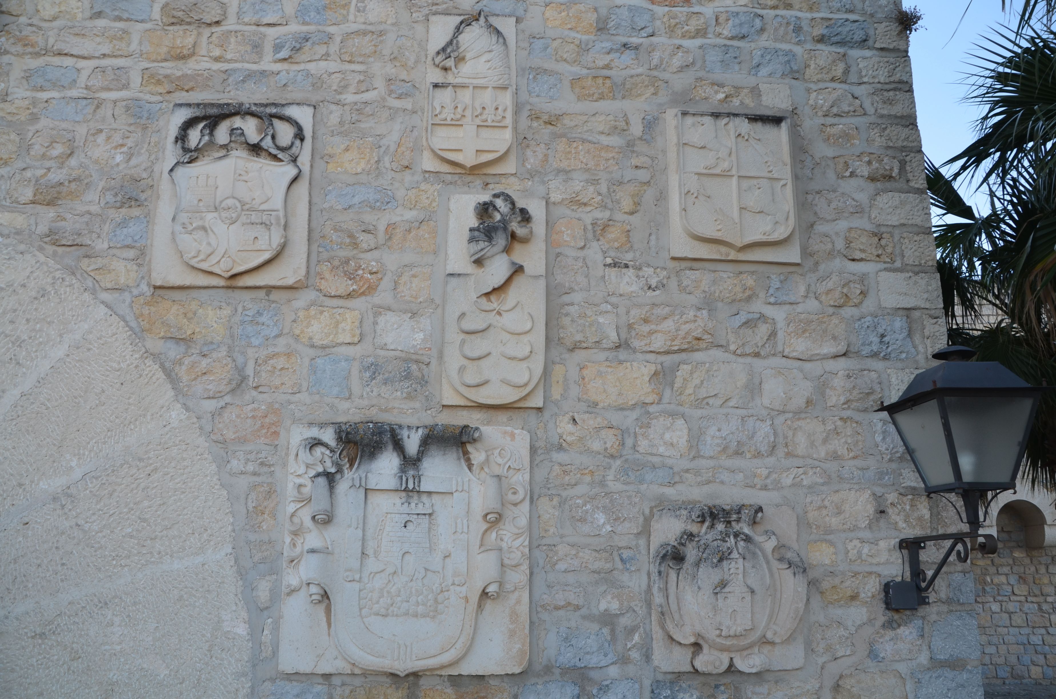 A stone wall with multiple coats of arms and a street lamp.