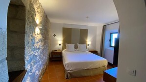 Un dormitorio con pared de piedra, una cama con sábanas blancas, dos mesitas de noche y luces fijadas en la pared.