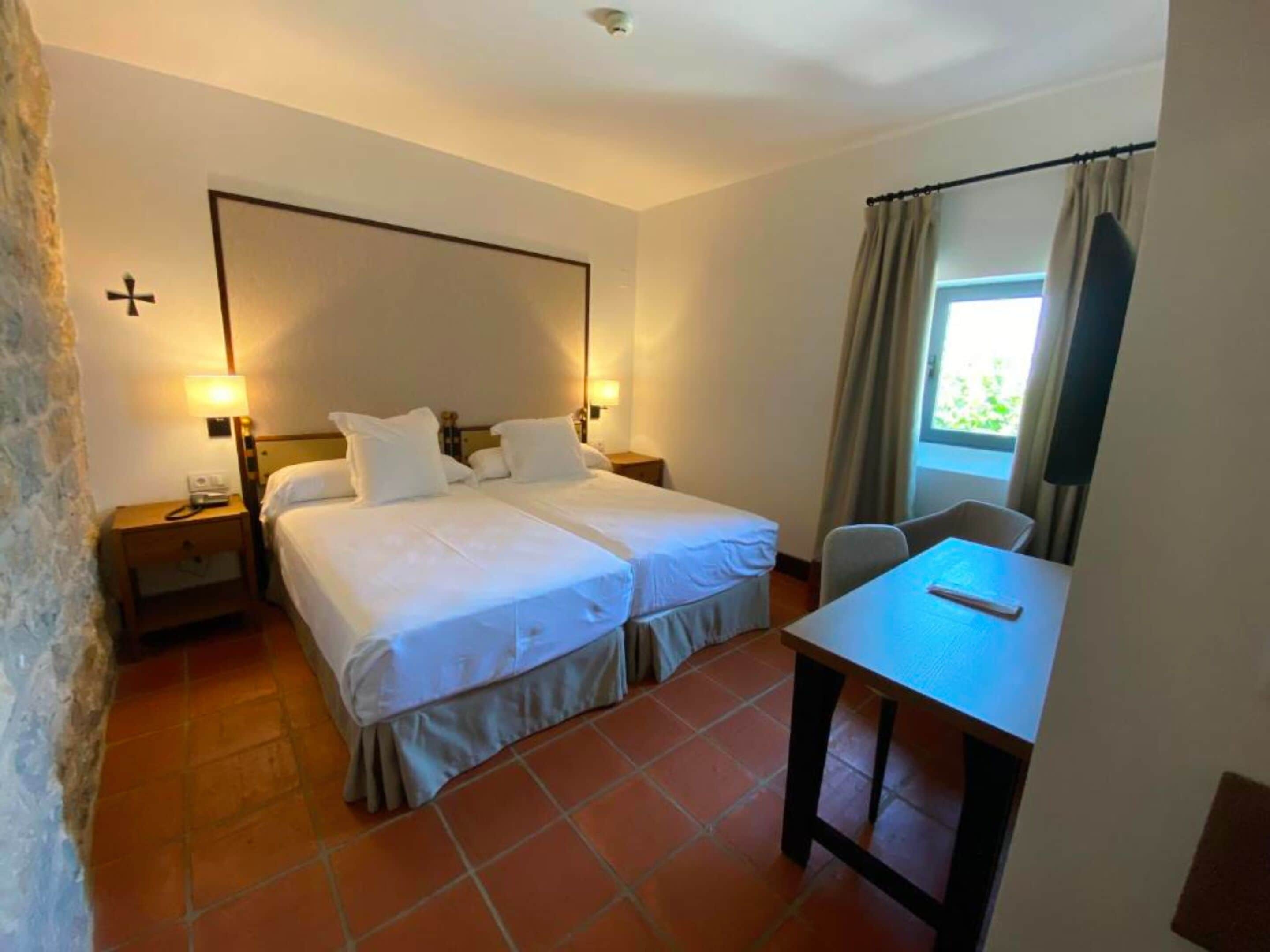 Une chambre d’hôtel avec deux lits, un bureau, une chaise, une fenêtre avec des rideaux et une croix au mur.