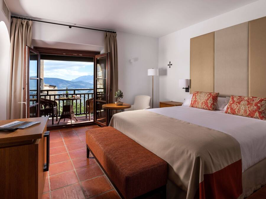 A hotel room with a large bed, a desk with a keyboard, a chair, a small table with a lamp, and a balcony with a view of mountains.