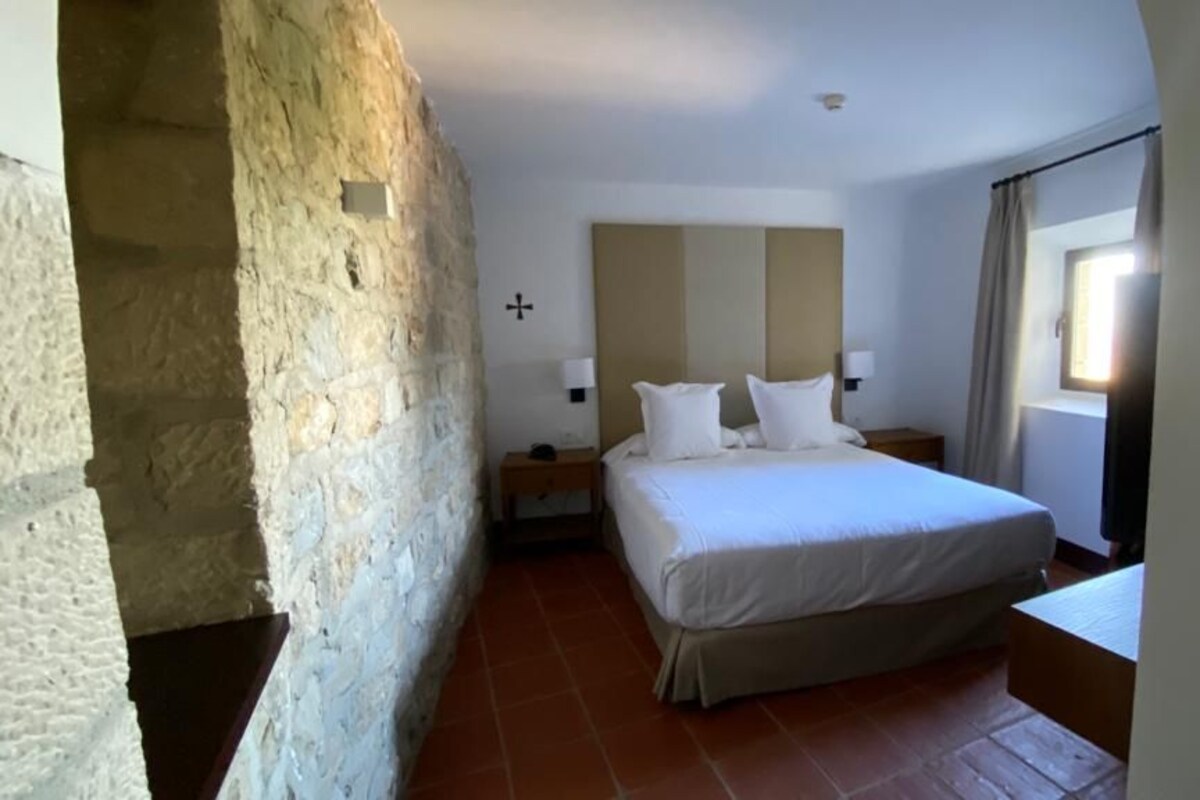 Une chambre à coucher avec un lit, des tables de chevet, un mur en pierre et une fenêtre avec des rideaux.