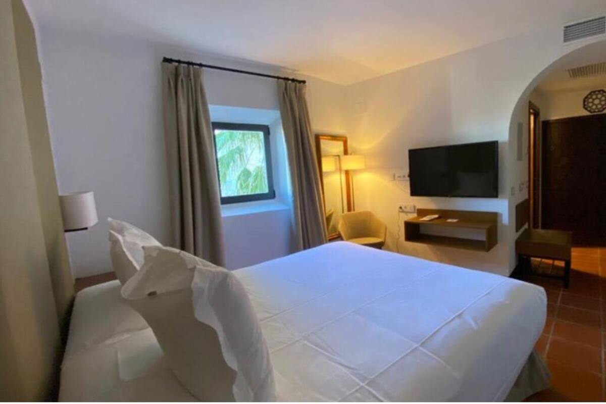 Una habitación de hotel con una cama grande, televisión, una silla y un ventanal con cortinas.