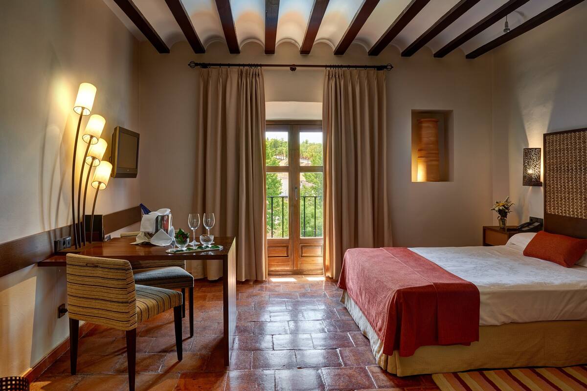 Ein Hotelzimmer mit einem Bett, einem Schreibtisch mit Stuhl, einem Tisch mit Lampe und Blick auf Grünfläche durch das Fenster.