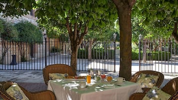 Mesa al aire libre con sillas de mimbre, mantel blanco y vista a una calle con valla y árboles.