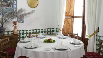 Mesa de jantar posta para quatro, com toalhas brancas, paredes com padrão verde e branco e um armário de madeira com uma panela de cobre.