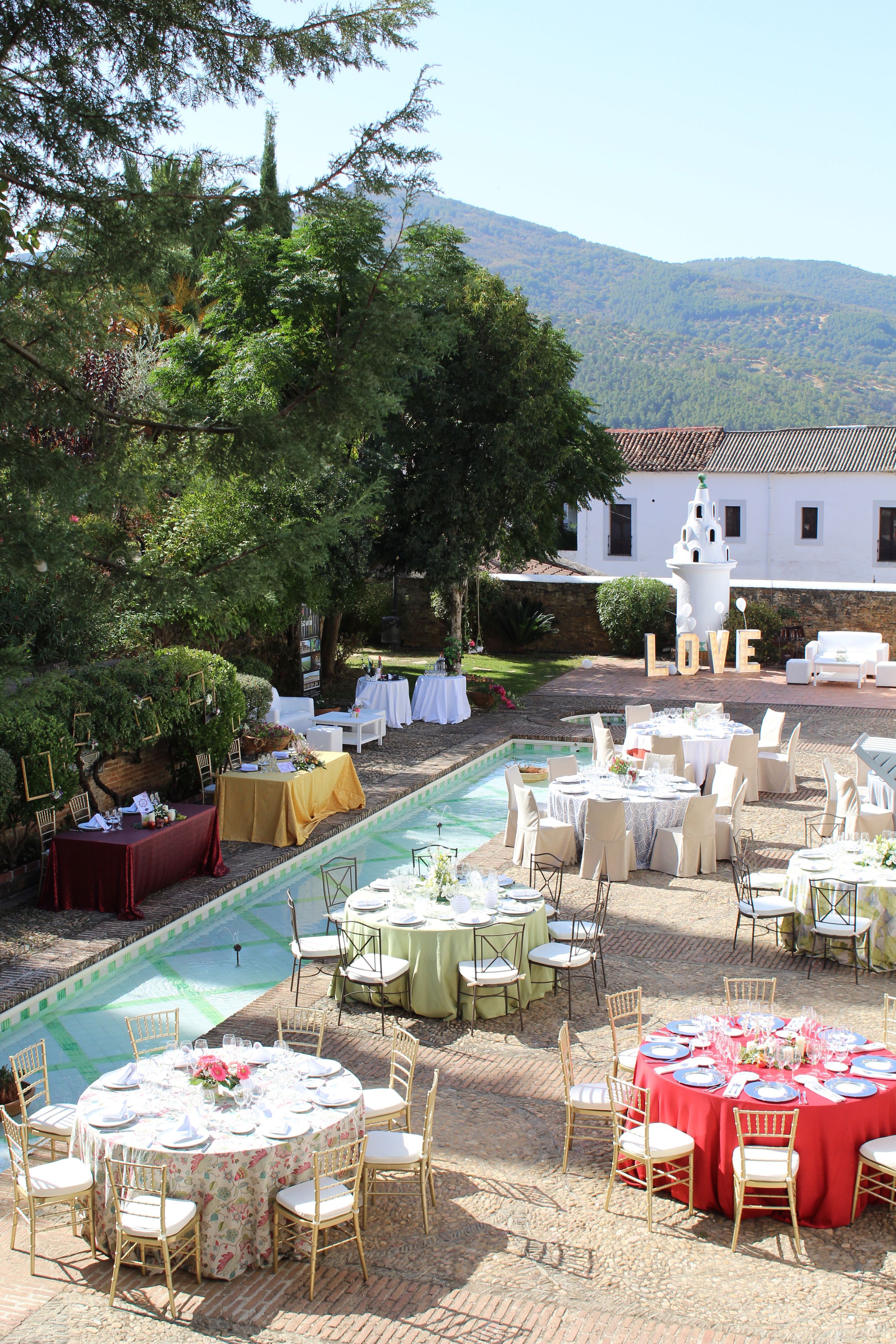 Un espace de restauration en plein air, avec des tables rondes dressées pour un repas, entouré d’une végétation luxuriante et d’une piscine.