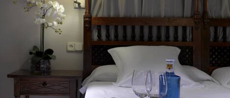 A hotel room with a bed, a bedside table with a lamp, a vase of white flowers, and a tray with teapot, cups, and a bottle.