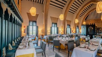 Un restaurante con techos altos en arco, varias mesas de comedor con manteles blancos y sillas, y lámparas colgantes.