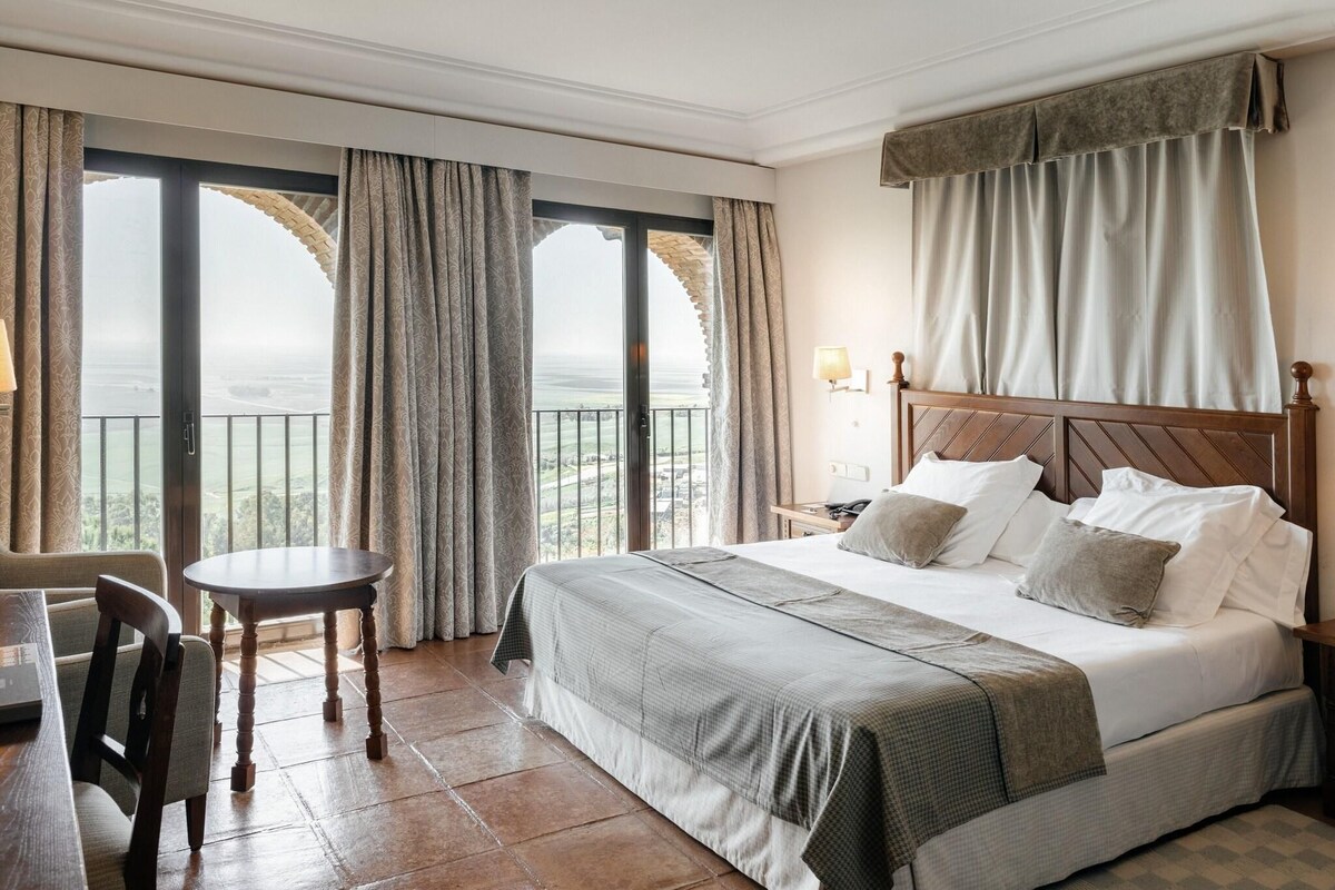 A hotel room with a large bed, a small table, and a chair. There is a balcony with curtains and a view of the ocean.