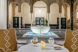 Un comedor con una mesa dispuesta para dos, con una vela y vasos de bebidas. Al fondo, una fuente grande y arcos.