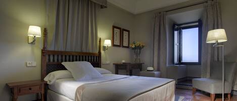A bedroom with a large bed, bedside lamps, a nightstand, a chair, a window, and a vase with flowers.