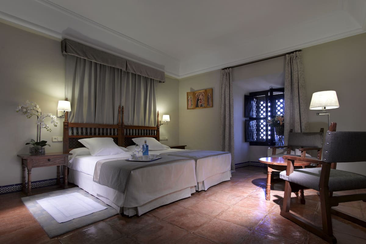 Une chambre d’hôtel avec deux lits, un bureau, une chaise, une fenêtre avec des rideaux et une table de chevet avec une lampe.