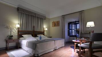 Quarto de hotel com duas camas, uma escrivaninha, uma cadeira, uma janela com cortinas e uma mesa de cabeceira com abajur.