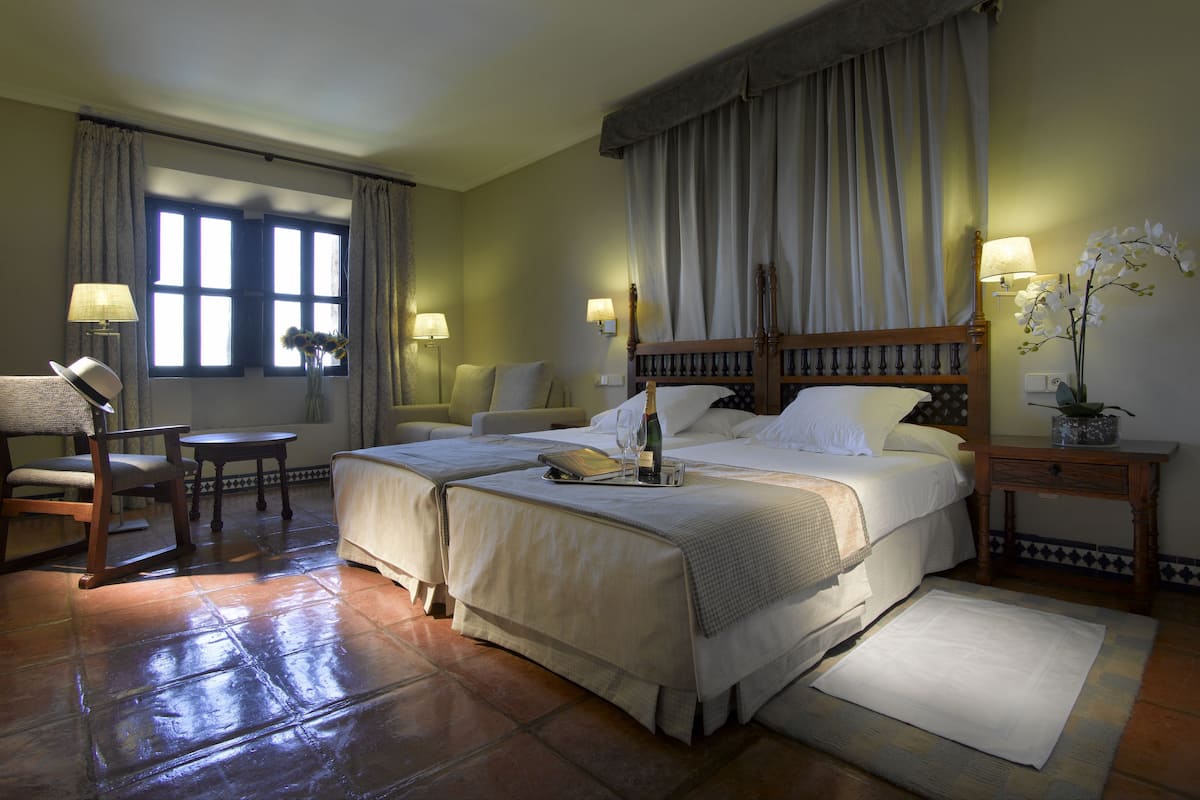 Une chambre d’hôtel avec un grand lit, deux tables de chevet, une chaise, une petite table et une fenêtre avec des rideaux.