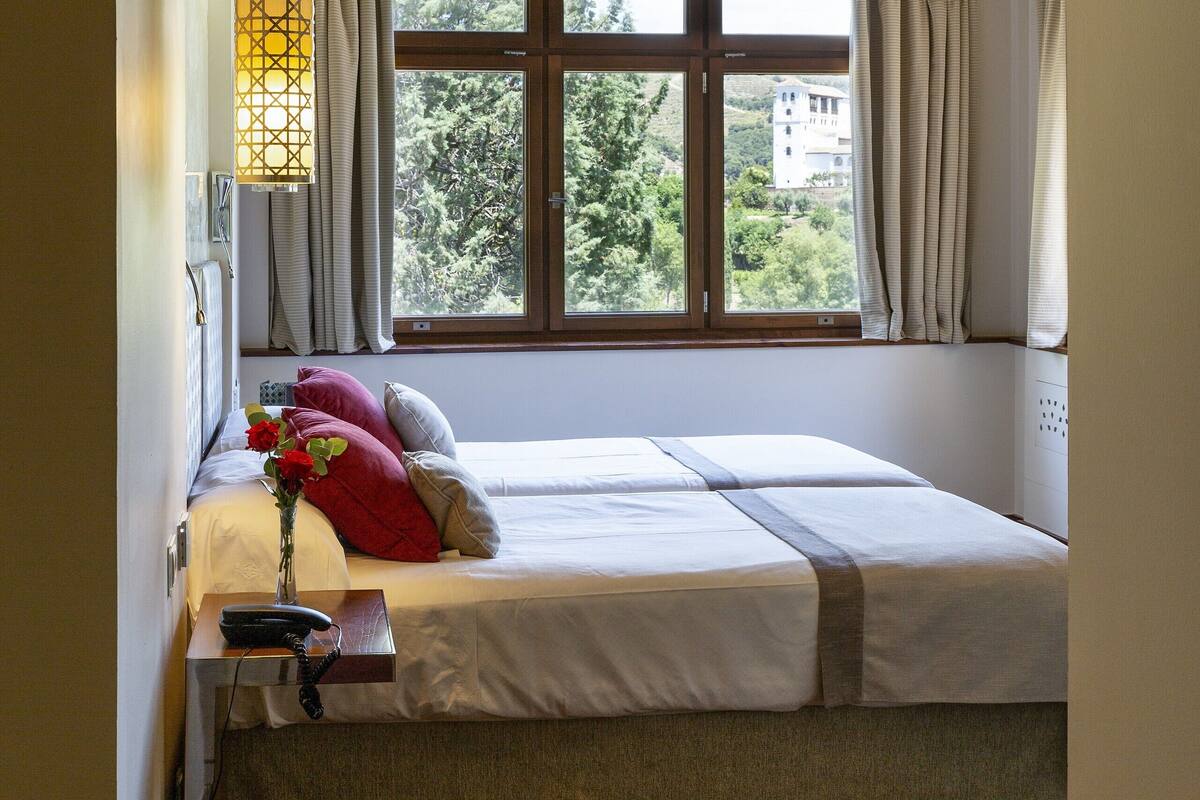 Quarto com teto de madeira, janela com cortinas, cama com travesseiros e mesa de cabeceira com abajur.
