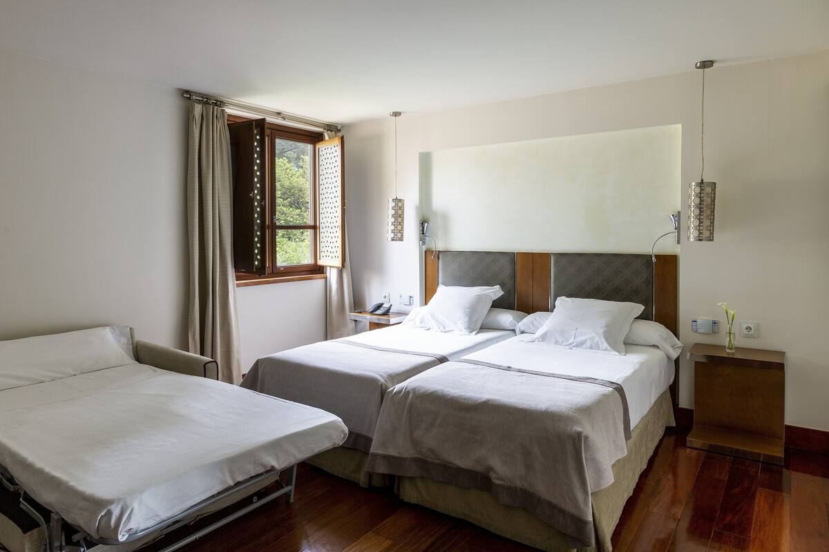 Quarto de hotel com duas camas de solteiro, uma mesa de cabeceira com abajur, uma janela com cortinas e um telefone ao lado da cama.
