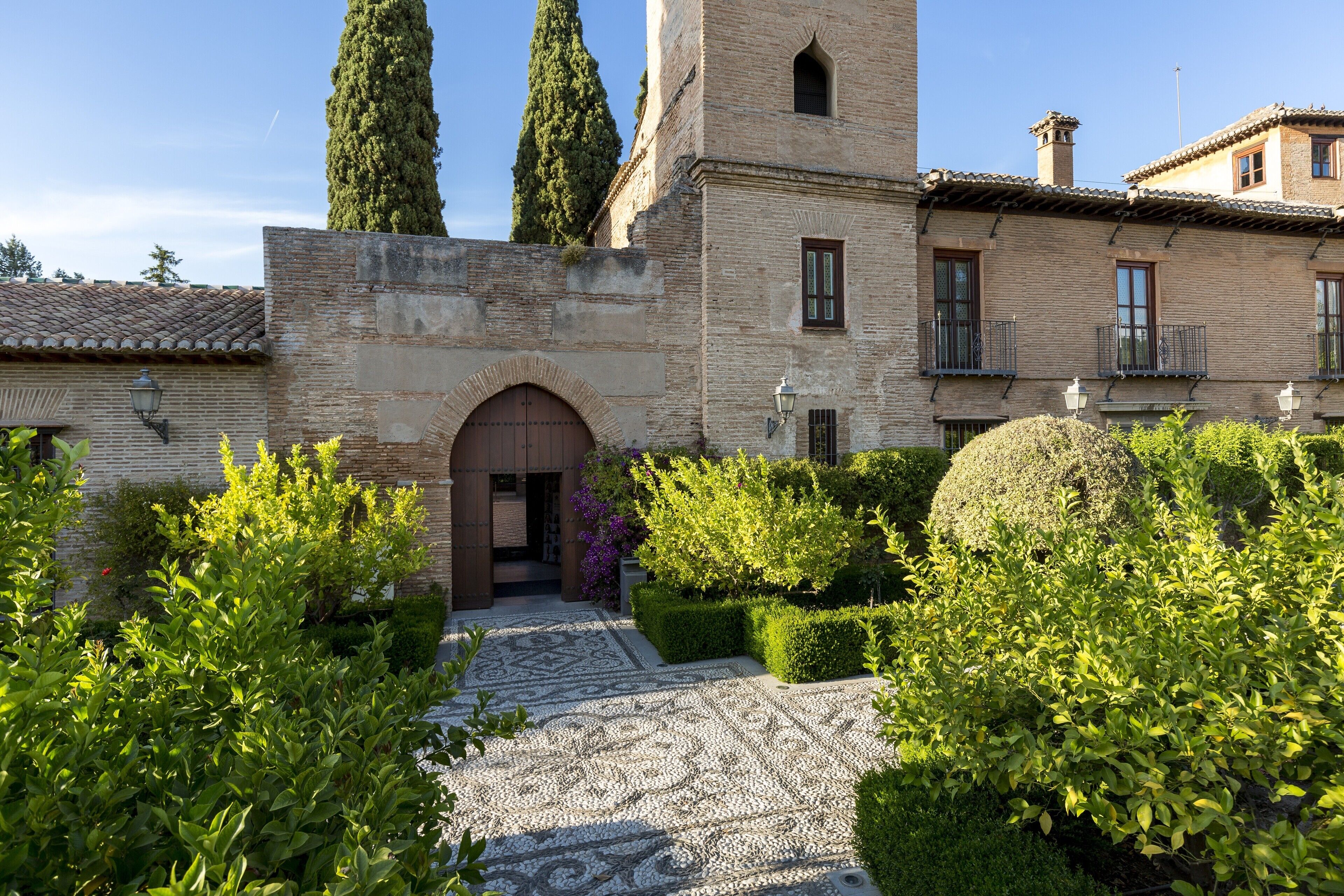 A courtyard with a patterned stone floor, a brick building with a tower, and neatly trimmed hedges.