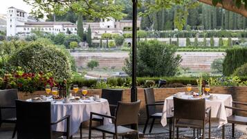 Un área de comedor al aire libre con mesas preparadas para dos, rodeada de follaje y vistas a un edificio histórico.