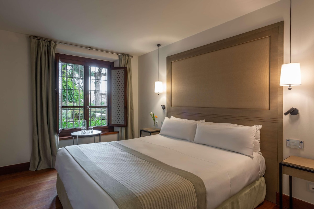A hotel room with a large bed, a small table, and a window with curtains.