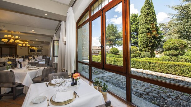 Un restaurante con mesas dispuestas para comer, ventanas amplias con vistas a un jardín y árboles.