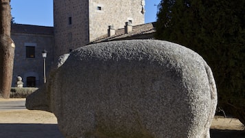 Una estatua de piedra de un animal, posiblemente un toro, en un patio con un edificio de piedra y un árbol al fondo.