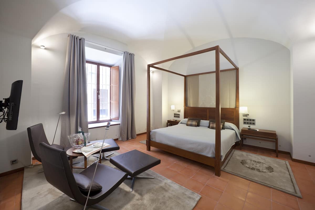 A bedroom with a four-poster bed, a desk with a chair, a TV, and a window with curtains.