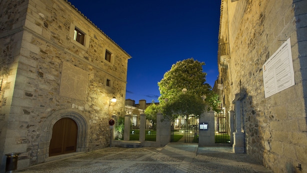 A historic stone building with a large wooden door, a paved courtyard, and a tree in the center.