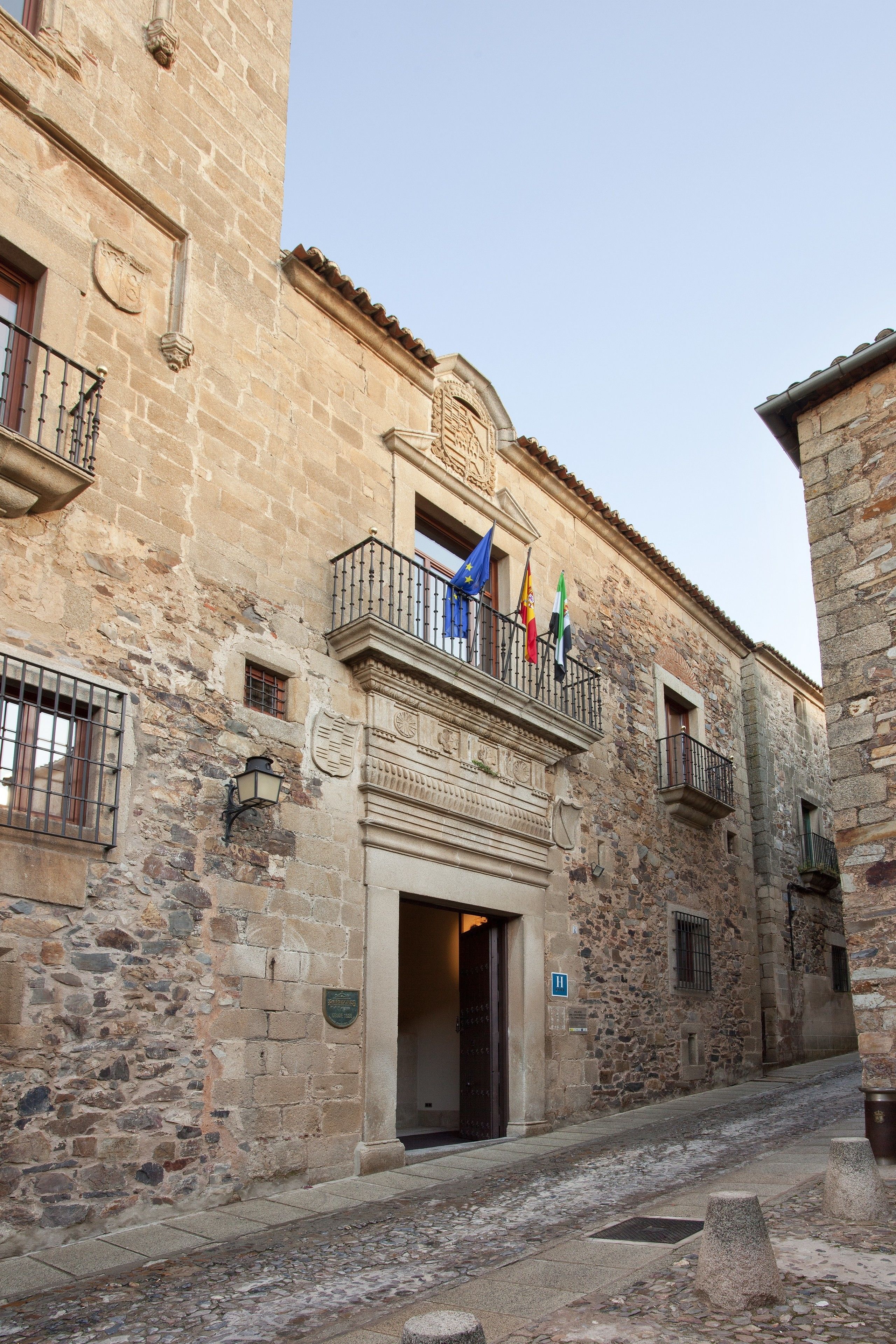 Un bâtiment historique en pierre, doté d’un balcon orné de drapeaux, une rue pavée et un sentier bordé de pierres.