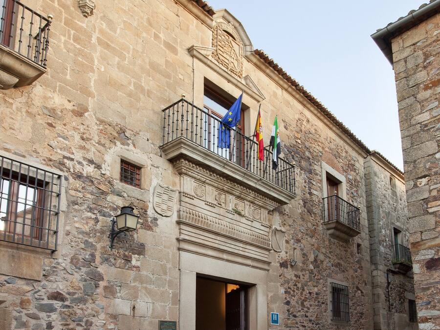 Un edificio histórico de piedra con un balcón que muestra banderas, una calle empedrada de piedra y un sendero adoquinado.