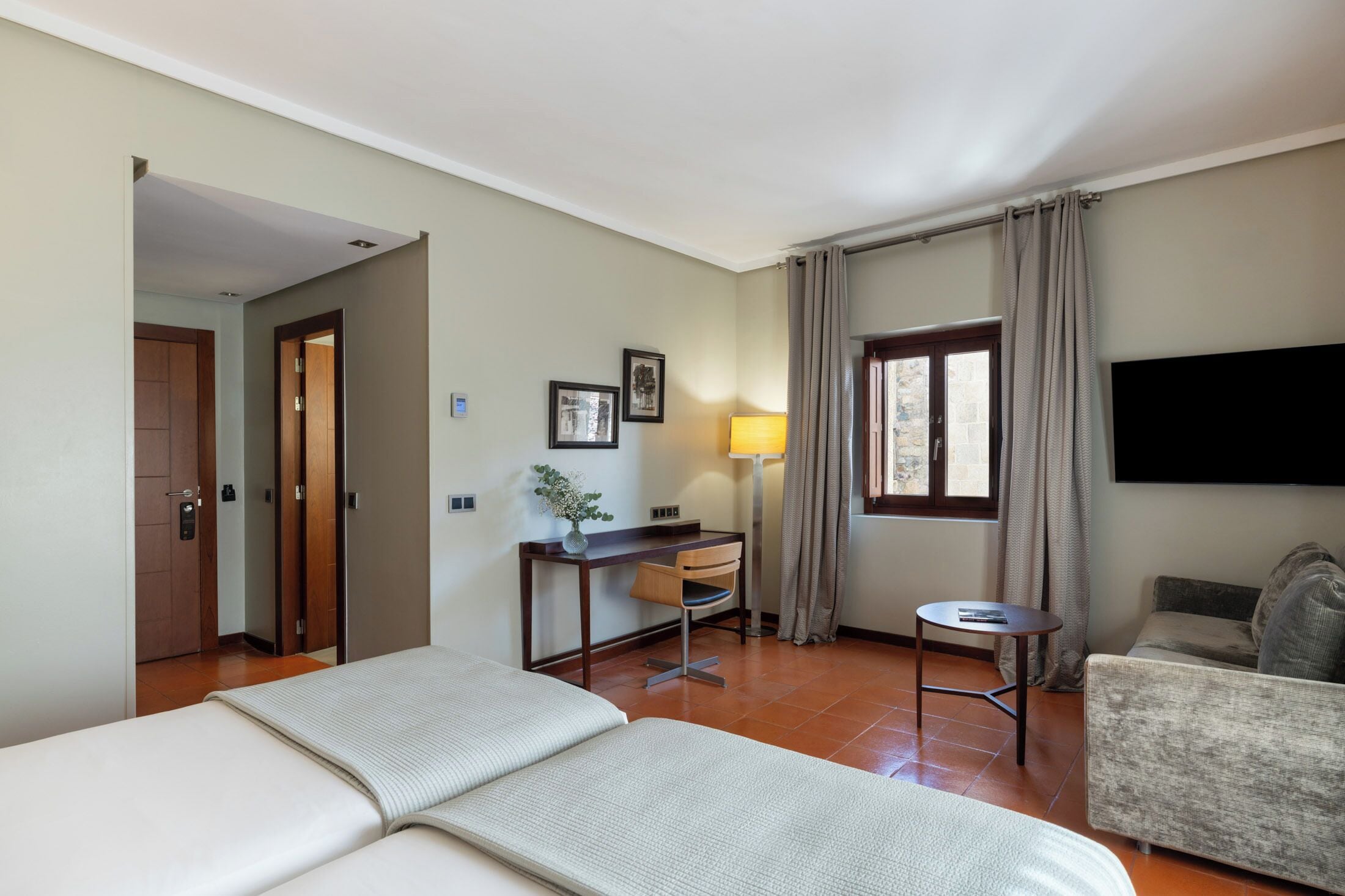 A hotel room with a bed, a desk, a chair, a sofa, a TV, and a window with curtains.