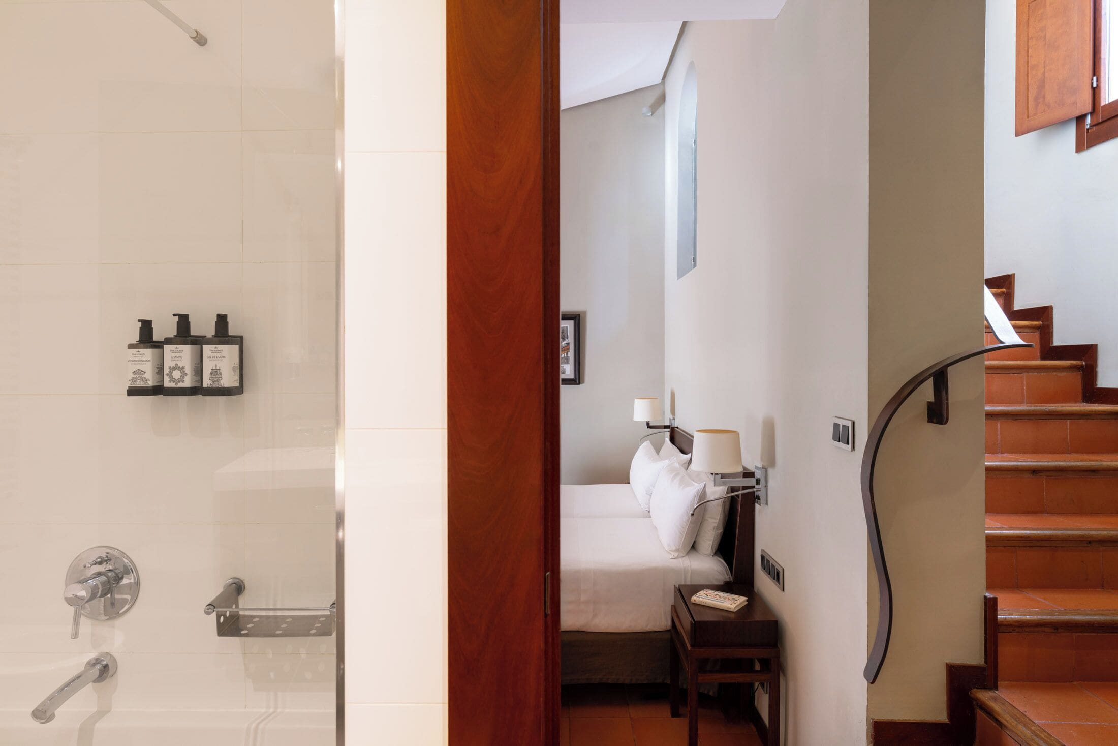 A bathroom with a glass shower enclosure, a wooden door, and a bed with a nightstand.