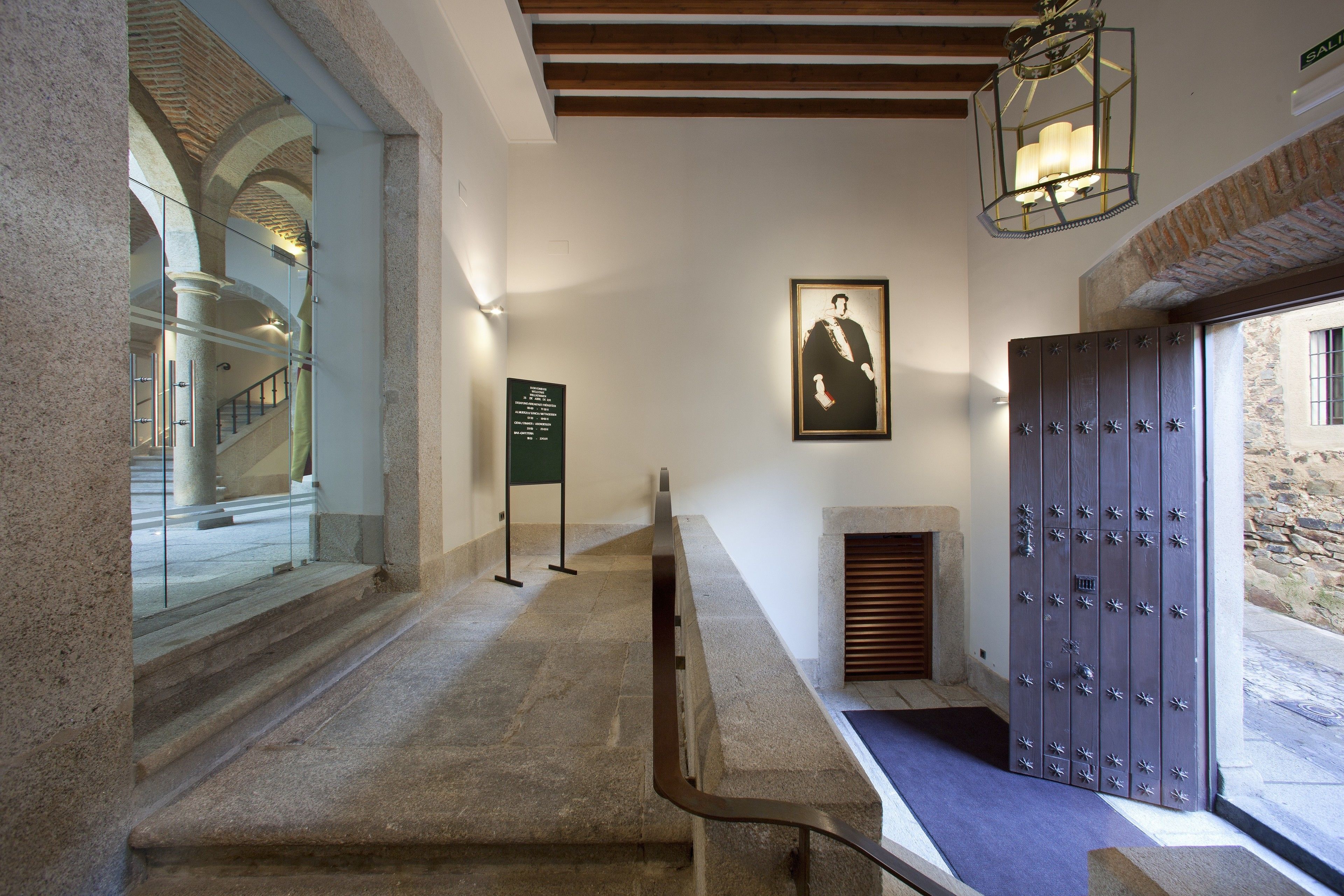 A hallway with stone steps, a framed portrait on the wall, a wooden door with metal studs, and a staircase visible through a glass door.