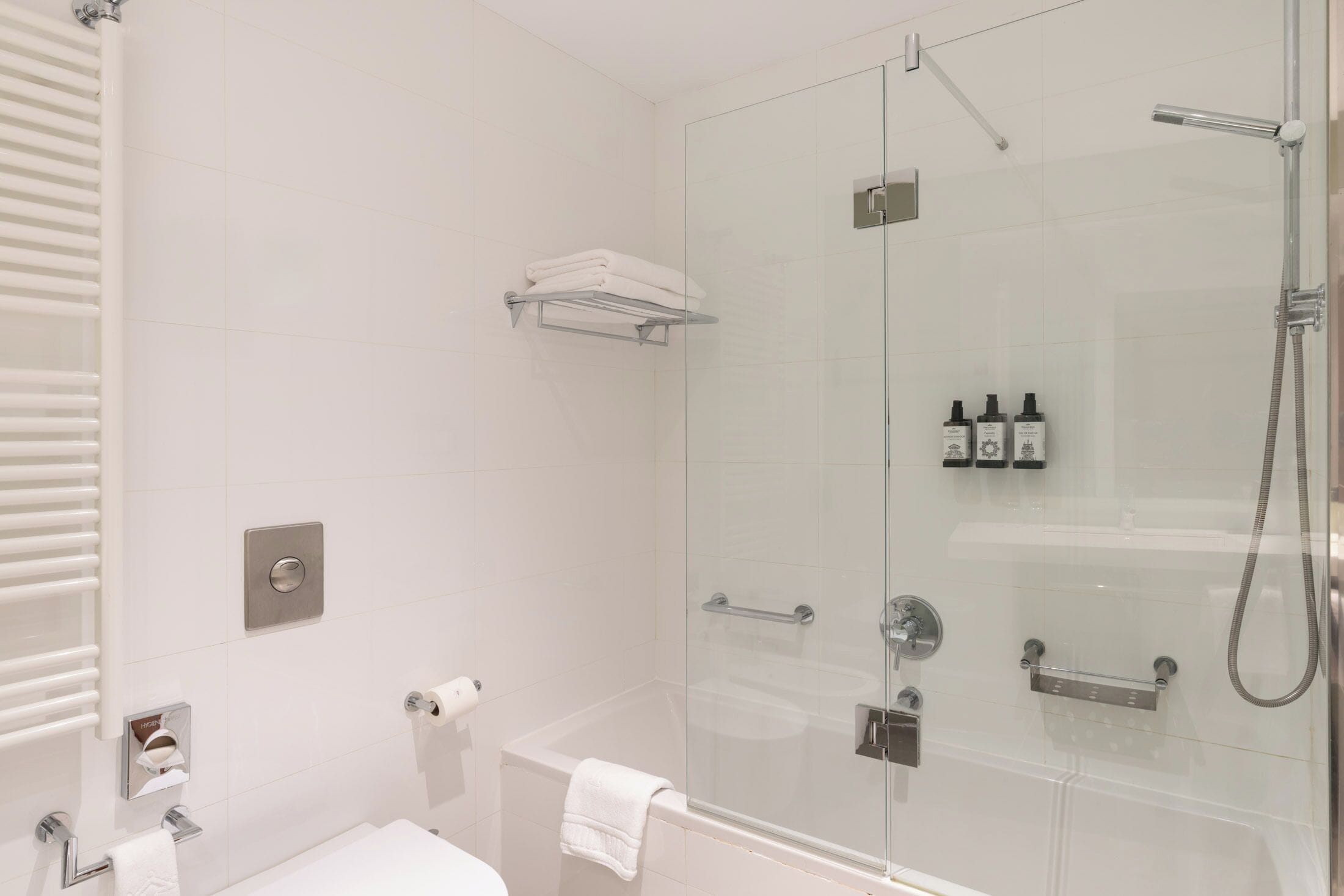 Un baño moderno con ducha de vidrio, inodoro blanco, barra para toallas y estante con artículos de aseo.